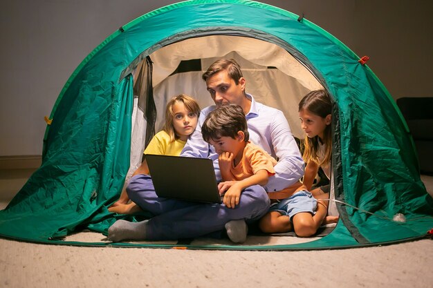 Schöne Kinder, die mit Vater im Zelt zu Hause entspannen und Film auf Laptop-Computer schauen. Nette Kinder und Vater mittleren Alters sitzen und haben Spaß zusammen. Kindheits-, Familienzeit- und Wochenendkonzept