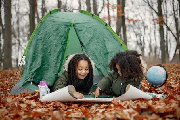 Schöne kleine schwarze Mädchen im Zeltcamping im Wald Zwei kleine Schwestern, die in einem Zelt im Herbstwald liegen und auf einer Karte suchen Schwarze Mädchen, die Khaki-Mäntel tragen