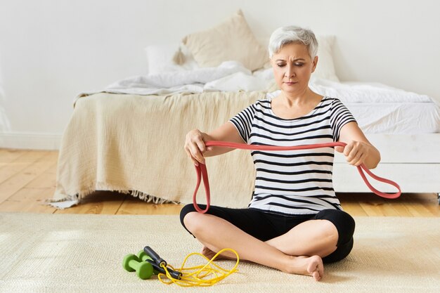 Schöne konzentrierte grauhaarige Rentnerin, die Übungen für Armmuskeln unter Verwendung des Gummibands macht und mit Springseil und Hanteln auf dem Boden sitzt. Alter, reife Menschen und aktiver Lebensstil