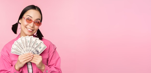 Schöne koreanerin mit sonnenbrille, die lächelndes zufriedenes konzept von schnellen krediten, mikrokrediten und zahlungen zeigt, die über rosa hintergrund stehen