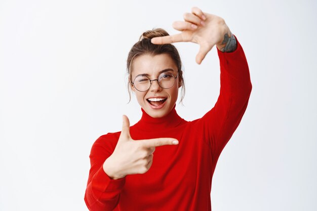 Schöne künstlerische Frau mit blonden Haaren und Brille, Handrahmenkamera machend und vorne zwinkernd, fotografieren, gegen weiße Wand stehend