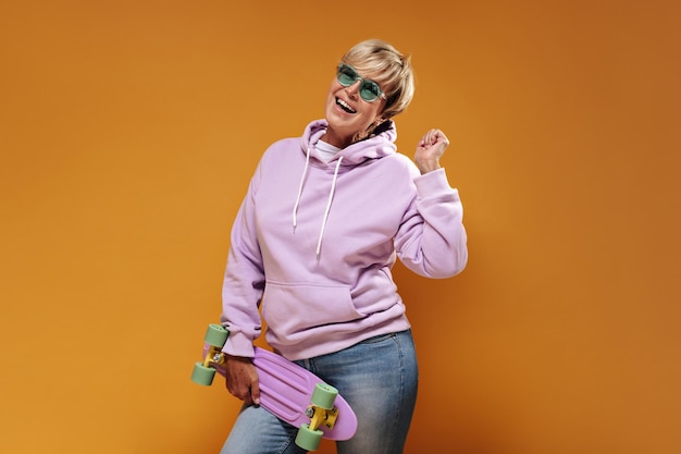Schöne kurzhaarige frau mit moderner sonnenbrille in rosafarbenem, coolem hoodie und blauer hose, die lacht und skateboard hält