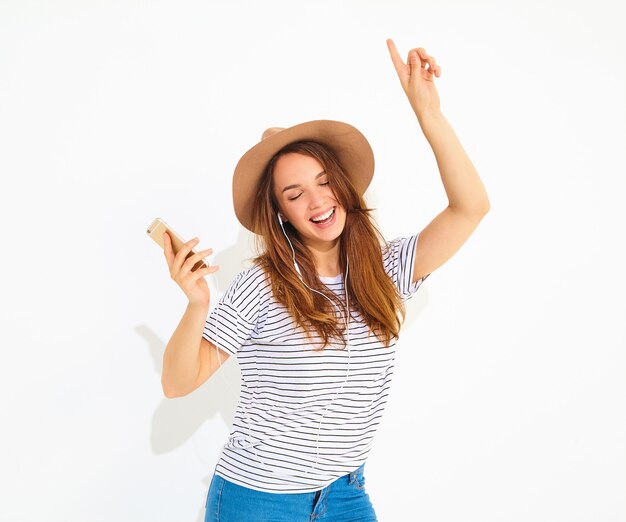 schöne lächelnde Brunettefrauenfrau im zufälligen Hippiesommer kleidet ohne Make-up in hörender Musik des braunen Hutes im Telefon mit den Kopfhörern, die auf Weiß lokalisiert werden