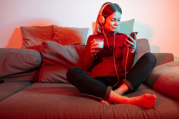 Kostenloses Foto schöne lächelnde dame mit kopfhörern, die zu hause auf dem sofa sitzt und musik mit einem glas milch und einem handy in den händen hört