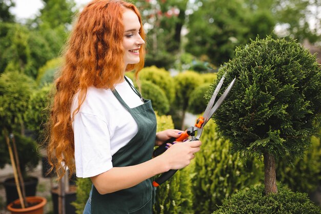 Schöne lächelnde Dame mit rothaarigem lockigem Haar, die in der Schürze steht und eine große Gartenschere hält, während sie im Freien arbeitet