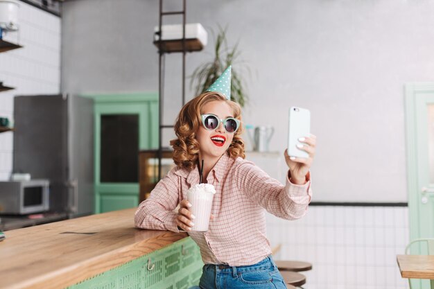 Schöne lächelnde Dame mit Sonnenbrille und Geburtstagskappe, die mit Milchshake an der Bartheke sitzt und süße Fotos auf ihrem Handy im Café macht