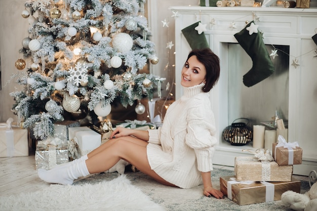 Schöne lächelnde Frau, die nahe Weihnachtsbaum mit Geschenken sitzt