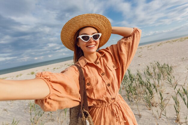 Schöne lächelnde Frau, die Selbstporträt macht und Ferien nahe dem Ozean genießt. Trägt eine trendige Retro-Sonnenbrille und einen Strohhut.