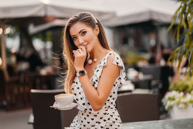 Schöne lächelnde Frau, die stilvolles weiß bedrucktes Kleid trägt, das im Straßencafé mit Tasse Cappuccino sitzt