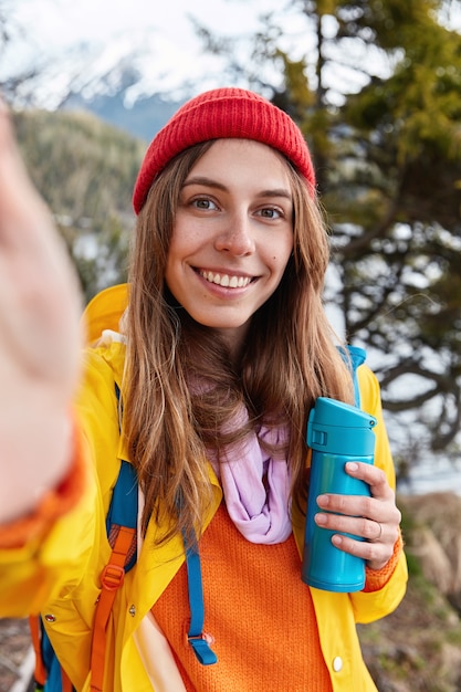 Schöne lächelnde Frau hält unerkennbares Gerät, macht Selfie, trägt roten Hut und gelbe Jacke