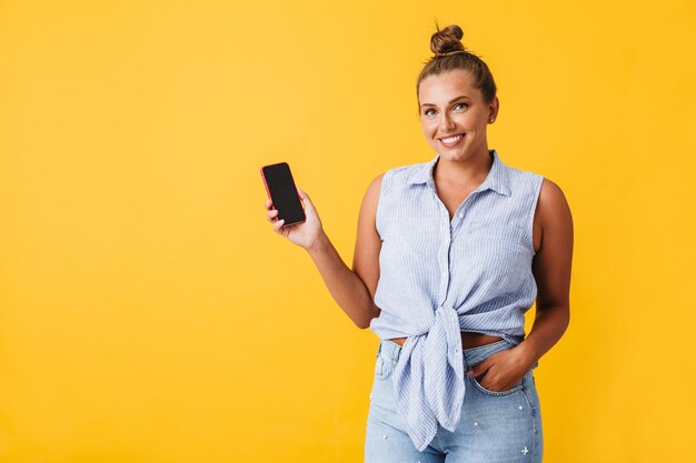 Schöne lächelnde Frau im Hemd, die glücklich in die Kamera schaut, während sie ein neues Mobiltelefon auf gelbem Hintergrund zeigt