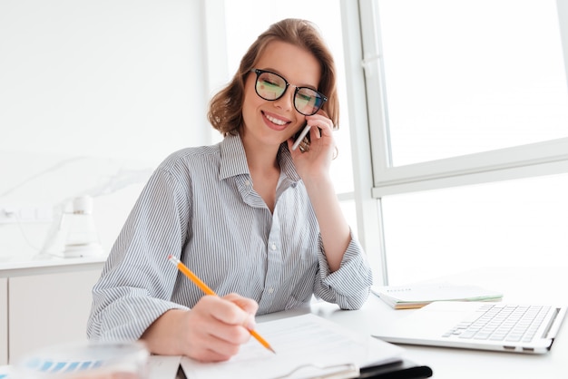 Schöne lächelnde Frau in den Gläsern, die auf Handy sprechen, während sie mit Dokumenten zu Hause arbeiten