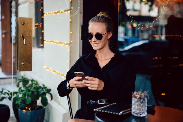 Schöne lächelnde Frau in der Sonnenbrille und im schwarzen eleganten Mantel, unter Verwendung eines Handys