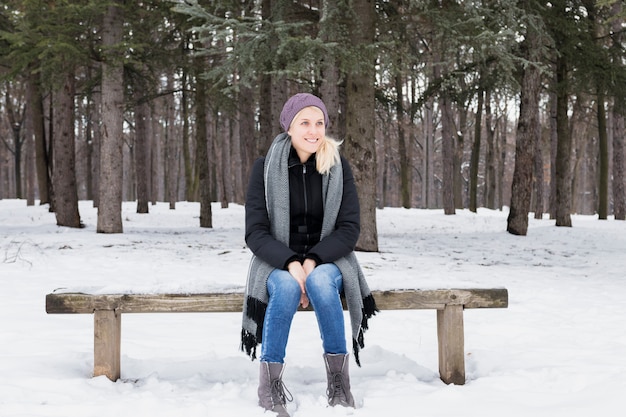 Schöne lächelnde junge Frau, die auf Holzbank im Winterwald sitzt