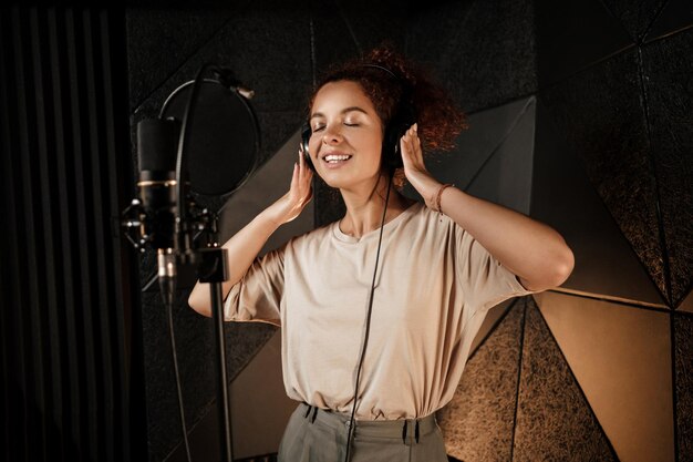 Schöne lächelnde Sängerin mit Kopfhörern, die träumerisch ein neues Lied im professionellen Studio aufnimmt