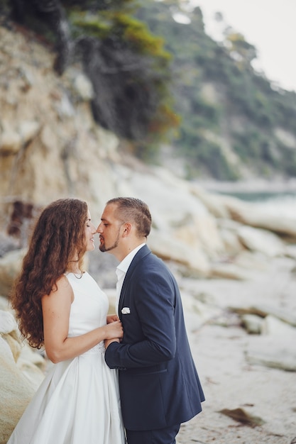 Schöne langhaarige Braut im weißen Kleid mit ihrem Ehemann auf dem Strand nahe großen Steinen
