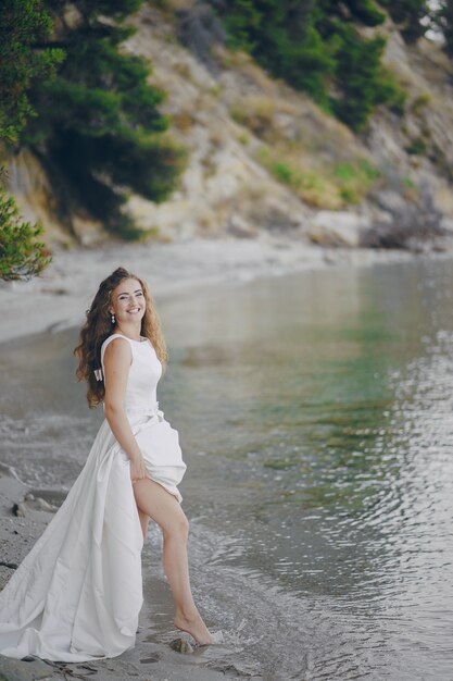 Schöne langhaarige Braut in einem ausgezeichneten weißen Kleid gehend auf einen Strand