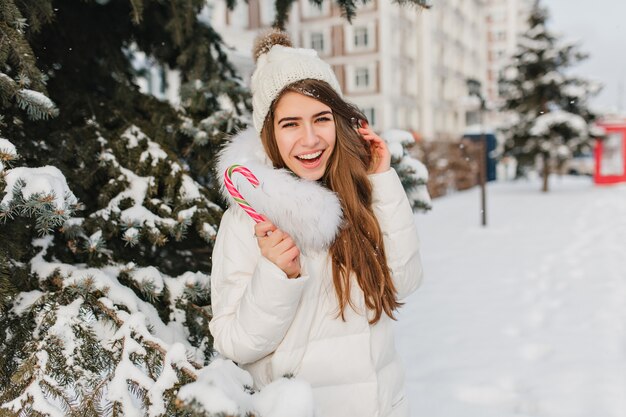 Schöne langhaarige Frau, die Winterferien genießt und mit leckerem Zuckerstange aufwirft. Außenporträt der inspirierten kaukasischen Dame im lustigen Hut, der auf Weihnachten wartet und auf der verschneiten Straße herumalbert.