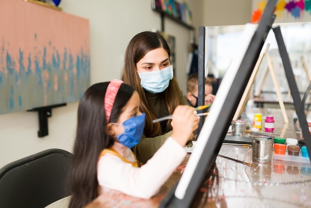 Schöne Lateinlehrerin und junges kaukasisches Mädchen mit Gesichtsmasken malen mit einem Pinsel auf einer leeren Leinwand für die Kunstschule