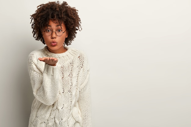 Schöne liebevolle afroamerikanische Frau bläst Luftkuss von offener Handfläche, drückt Hingabe aus, trägt runde Brille und weißen Pullover, posiert mit Leerzeichen für Ihre Informationen.