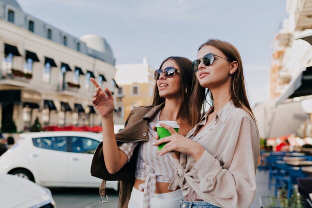 Schöne Mädchen in Sonnenbrillen halten Kaffee, benutzen ein Smartphone und lächeln, während sie draußen stehen.