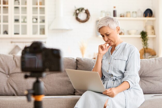 Schöne moderne ältere grauhaarige Frau in stilvollen Kleidern, die entfernt mit Laptop arbeiten, Nachricht tippen, nachdenklichen Gesichtsausdruck haben, auf Sofa sitzen