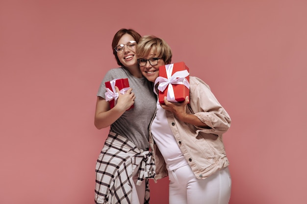 Schöne moderne zwei Frauen mit kurzer Frisur und Brille in der weißen Hose lächelnd, umarmt und hält rote Geschenkboxen auf rosa Hintergrund.