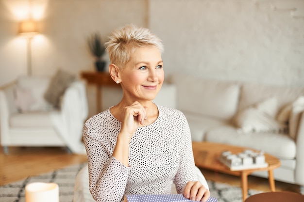 Schöne modische europäische frau in ihren fünfzigern, die den blick absorbiert, während sie neujahrsgeschenke einpackt, über glückwünsche und wünsche nachdenkt und glücklich lächelt. urlaub und festliche stimmung