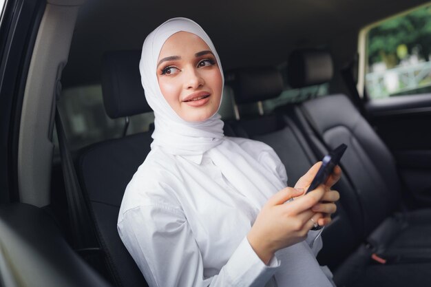 Schöne muslimische Frau mit weißem Hijab, die auf dem Rücksitz eines Autos sitzt und Smartphone benutzt