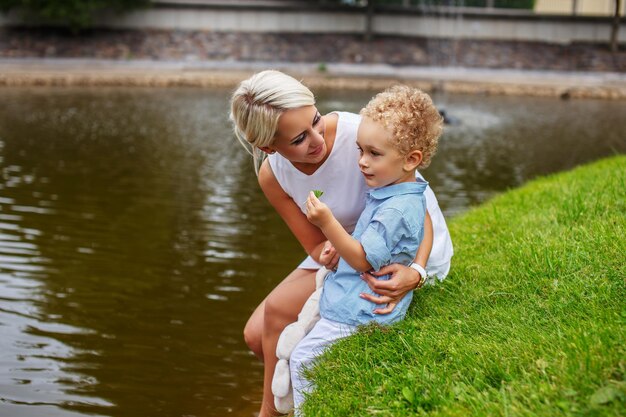 Schöne Mutter mit ihrem Kind, das auf Gras in der Nähe des Flusses sitzt.