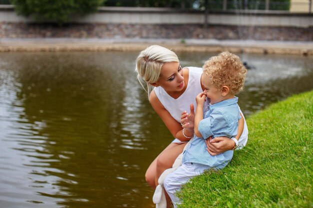 Schöne Mutter mit ihrem Kind, das auf Gras in der Nähe des Flusses sitzt.