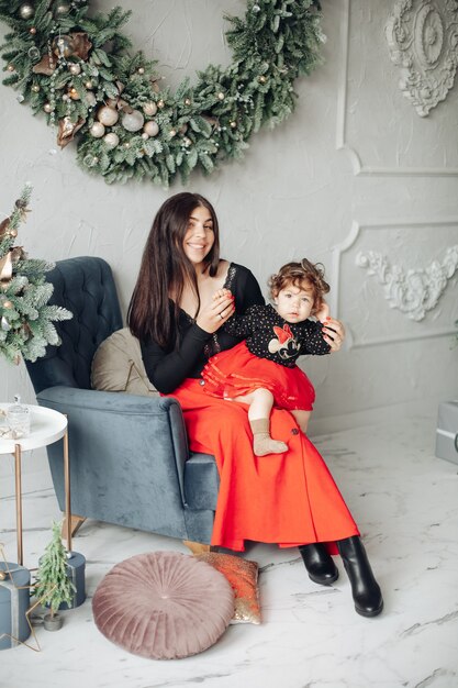 Schöne Mutter und ihre süße kleine Tochter sitzen in einem Sessel unter Weihnachtskranz