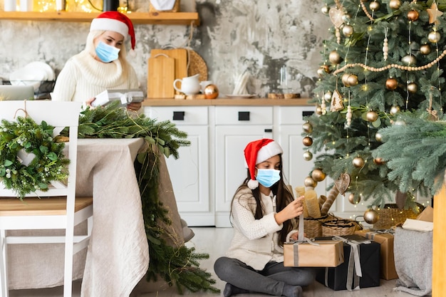 Schöne Mutter und Tochter in medizinischen Masken haben Spaß zu Hause in der Nähe des Weihnachtsbaums in einem Innenraum. Familienglück, Urlaub, Freude, Urlaub, Spiele mit einer Frau. Neujahrsvorbereitungen.