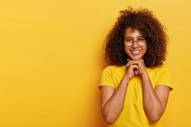 Schöne positive Afro-Frau hat lockiges Haar, gesunde Haut, hält die Hände unter dem Kinn zusammen, freut sich über angenehme Kommentare zu ihrer Arbeit, trägt ein gelbes T-Shirt, Modelle im Innenbereich. Konzept der menschlichen Emotionen