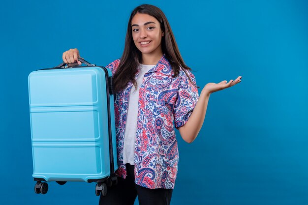 Schöne Reisende Frau, die mit Reisekoffer mit Lächeln auf Gesicht steht, das mit Arm der Hand über isoliertem blauem Raum präsentiert