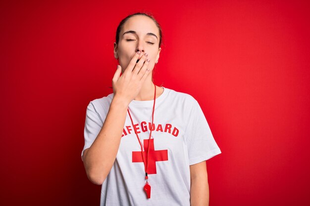 Schöne Rettungsschwimmerin trägt T-Shirt mit rotem Kreuz und pfeift vor isoliertem Hintergrund, gelangweilt, gähnt, müde und bedeckt den Mund mit der Hand. Unruhig und schläfrig