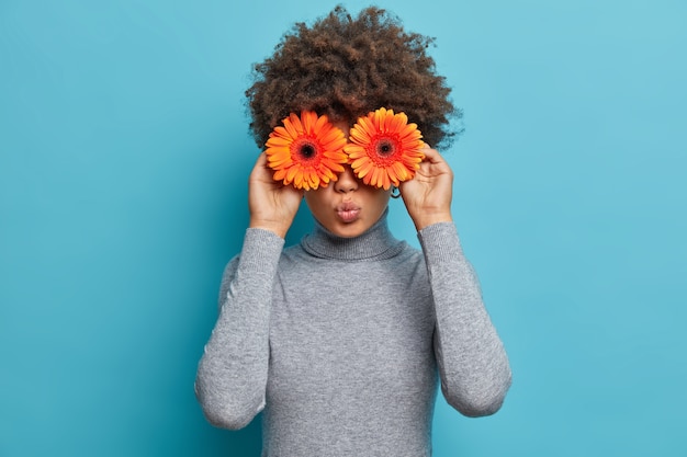 Schöne romantische junge frau mit blumen vor den augen, hält die lippen gerundet, hält orange gerbera gänseblümchen