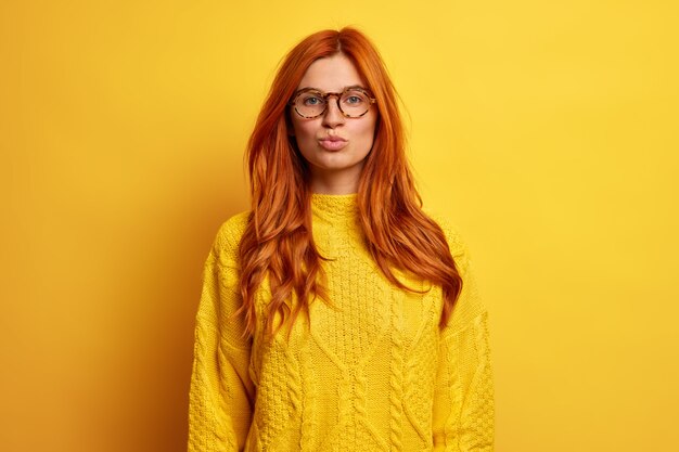 Schöne rothaarige Frau hält die Lippen gefaltet, als sie jemanden küssen will, der mit flirtendem Ausdruck aussieht, trägt eine transparente Brille und einen gestrickten Pullover.