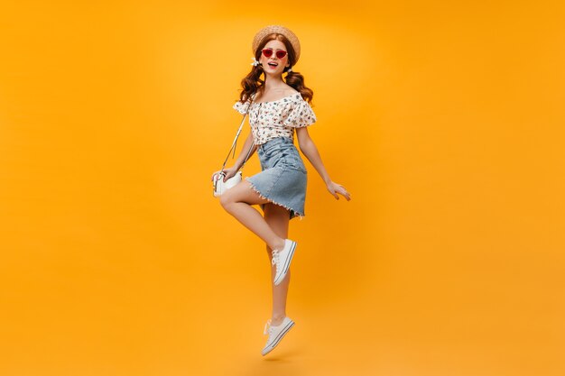 Schöne rothaarige Frau im Sommer-Denim-Outfit und im Strohhut, die weiße Tasche halten und auf orange Hintergrund springen.