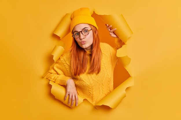 Schöne rothaarige junge Frau mit runden Lippen gekleidet in Hut und Pullover hat Flirtausdruck gekleidet in gelbe Kleidung steht durch zerrissenes Papier