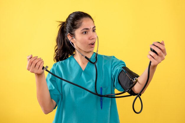 Schöne Ärztin der Vorderansicht in der Uniform, die Blutdruckmessgeräte hält, die auf gelbem Hintergrund stehen