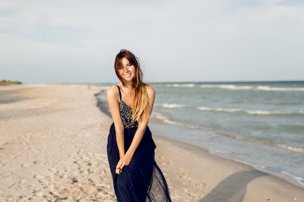Schöne schlanke Frau im blauen eleganten Kleid, das am sonnigen Strand nahe Ozean aufwirft. Windige Haare. Perfektes offenes Lächeln.