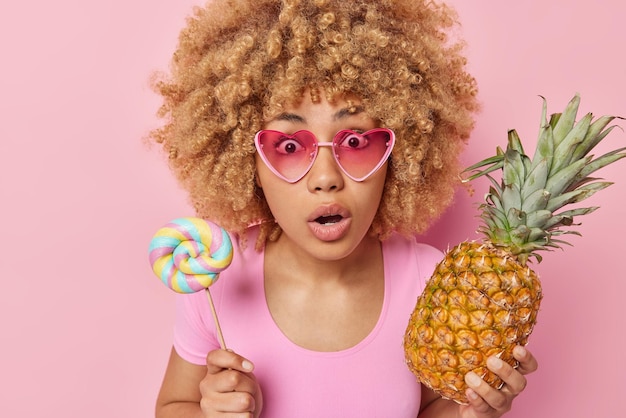 Schöne schockierte Frau mit lockigem Haar trägt herzrosa Sonnenbrille hält Lollipop und Ananas wählt zwischen gesundem und ungesundem Essen starrt ängstlich in die Kamera isoliert über rosa Hintergrund