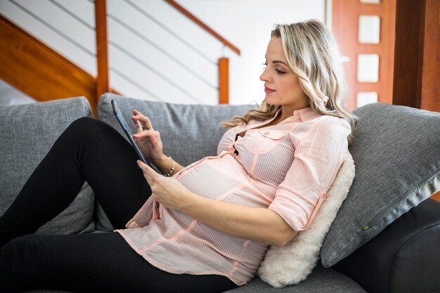 Schöne schwangere Frau, die digitale Tablette auf Sofa verwendet