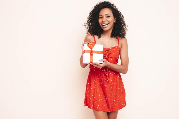 Schöne schwarze Frau mit Afro-Locken-Frisur Lächelndes Modell in rotem Sommerkleid gekleidet Sexy sorglose Frau posiert in der Nähe einer weißen Wand im Studio Gebräunt und fröhlich Holding-Geschenkbox