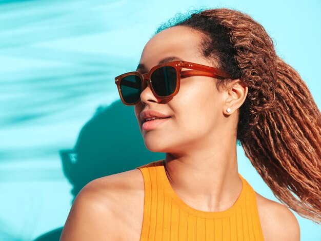 Schöne schwarze Frau mit Afro-Locken-Frisur Lächelndes Modell in Sommerkleidung gekleidet Sexy sorglose Frau posiert in der Nähe der blauen Wand im Studio Gebräunt und fröhlich Mit Sonnenbrille