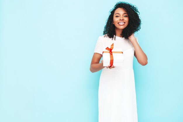 Schöne schwarze Frau mit Afro-Locken-Frisur Lächelndes Modell in weißem Sommerkleid gekleidet Sexy sorglose Frau posiert in der Nähe der blauen Wand im Studio Gebräunt und fröhlich Holding-Geschenkbox Isoliert