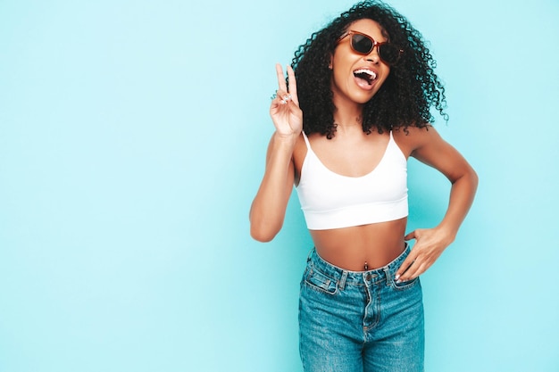 Schöne schwarze Frau mit Afro-Locken Frisur Lächelndes Modell in weißem Sommerkleid gekleidet Sexy sorglose Frau posiert in der Nähe der blauen Wand im Studio Gebräunt und fröhlich Isoliert In Sonnenbrille