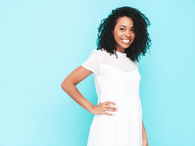 Schöne schwarze Frau mit Afro-Locken-Frisur Lächelndes Modell in weißem Sommerkleid gekleidet Sexy sorglose Frau posiert in der Nähe der blauen Wand im Studio Gebräunt und fröhlich Isoliert