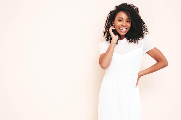 Schöne schwarze Frau mit Afro-Locken-Frisur Lächelndes Modell in weißem Sommerkleid gekleidet Sexy sorglose Frau posiert in der Nähe der Wand im Studio Gebräunt und fröhlich Isoliert